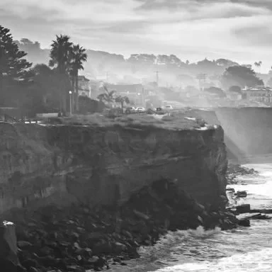 A coastal cliff scene in black and white. The rocky cliffs meet the ocean waves below, with palm trees and buildings visible in the distance. The atmosphere is misty and serene.