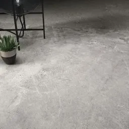 A close-up view of a concrete-like floor tile in a light grey shade, featuring subtle textures and variations. The surface has a matte finish, enhancing the tile's contemporary and industrial aesthetic. A potted plant and a modern chair are partially visible, adding to the minimalist design.