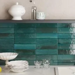 A kitchen backsplash with glossy, dark green rectangular tiles, neatly arranged in a horizontal stack bond pattern. The tiles have a rich, reflective finish that adds depth and a modern touch to the kitchen. The countertop below holds white dishes and glassware, creating a stylish contrast against the green tiles.