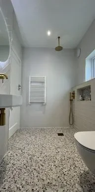 Image of a modern bathroom with white vertical striped tiles on the walls and a round illuminated mirror above a wooden vanity unit. The flooring consists of large beige tiles.