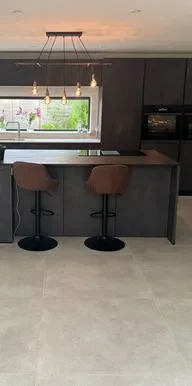 Modern kitchen featuring a sleek dark island with two brown bar stools, highlighted by a stylish light fixture. The space includes elegant floor tiles that seamlessly complement the contemporary design. 